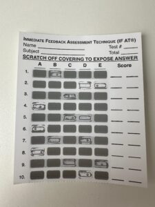 A photo of a completed Immediate Feedback Assessment Technique Sheet, showing some of the squares scratched off. An incorrect answer reveals a blank box, while a correct answer has a star in the box.