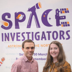 A photograph of Vicky Fawcett and a colleague, standing in front of a Space Investigators Poster